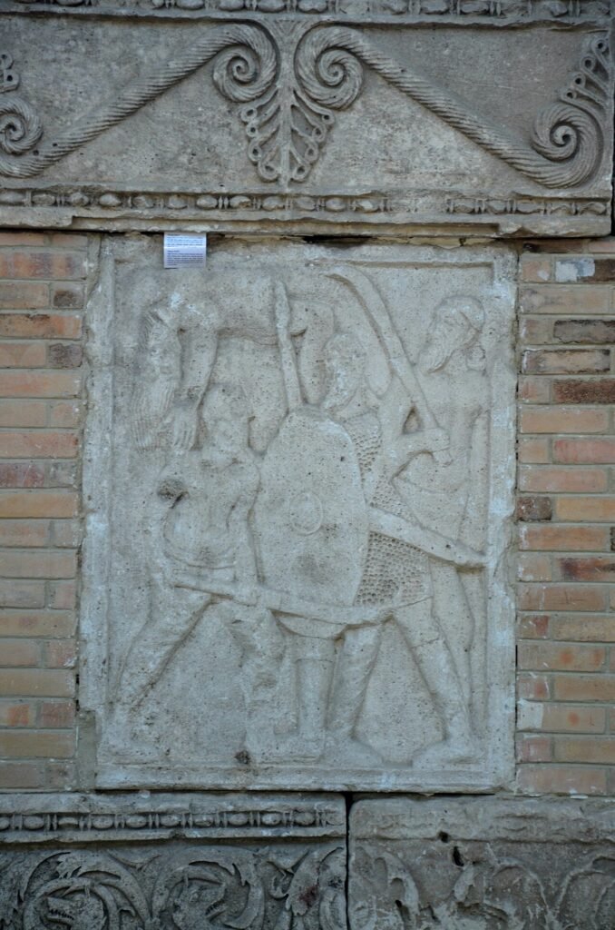 Metopă de calcar de la monumentul Tropaeum Traiani: soldat roman cu coif și platoșă luptând cu doi daci cu barbă, în pantaloni