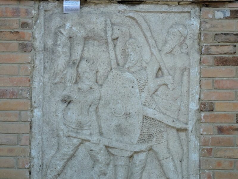 Metopă de calcar de la monumentul Tropaeum Traiani: soldat roman cu coif și platoșă luptând cu doi daci cu barbă, în pantaloni
