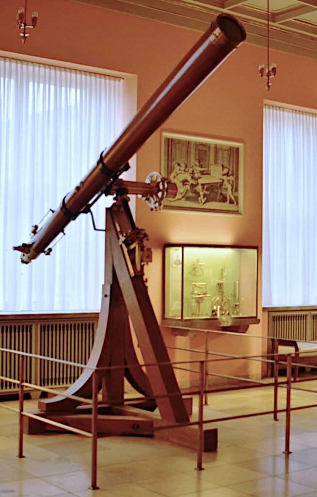 Telescop refractor istoric cu tub lung, montat pe un suport metalic, într-o sală de muzeu