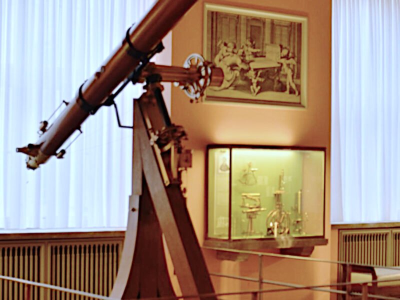 Telescop refractor istoric cu tub lung, montat pe un suport metalic, într-o sală de muzeu