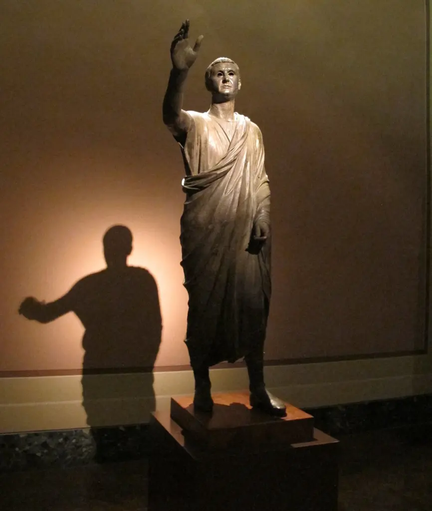 Statuie de bronz a unui bărbat în togă, cu brațul drept ridicat, expusă într-o sală de muzeu
